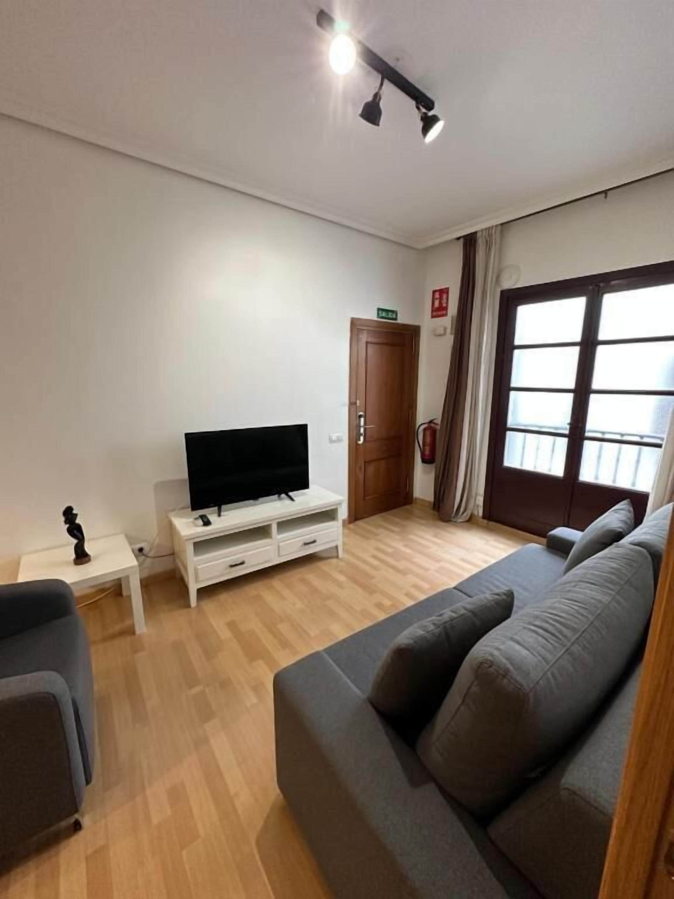 Un salón con un sofá gris, un televisor sobre un mueble blanco, una mesita con una estatua, y una puerta de madera con un letrero encima.