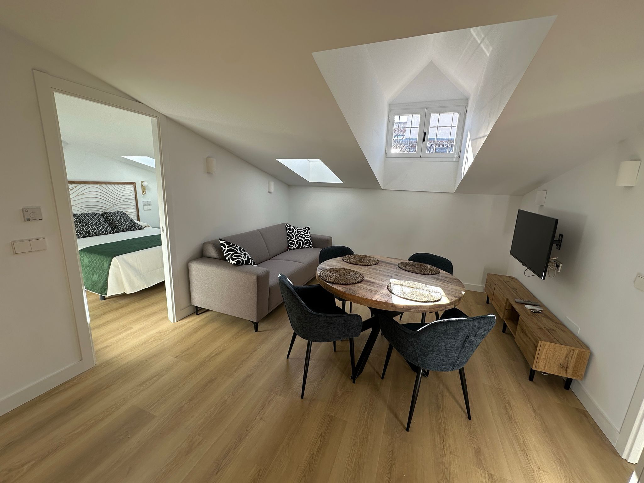 Un espacio habitable compacto con un entrepiso para dormir, una zona de comedor con mesa y sillas de madera, y un televisor instalado en la pared.