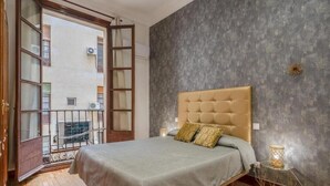 Un dormitorio con cama, una mesita de noche con lámpara, una ventana con vistas a edificios y un balcón.