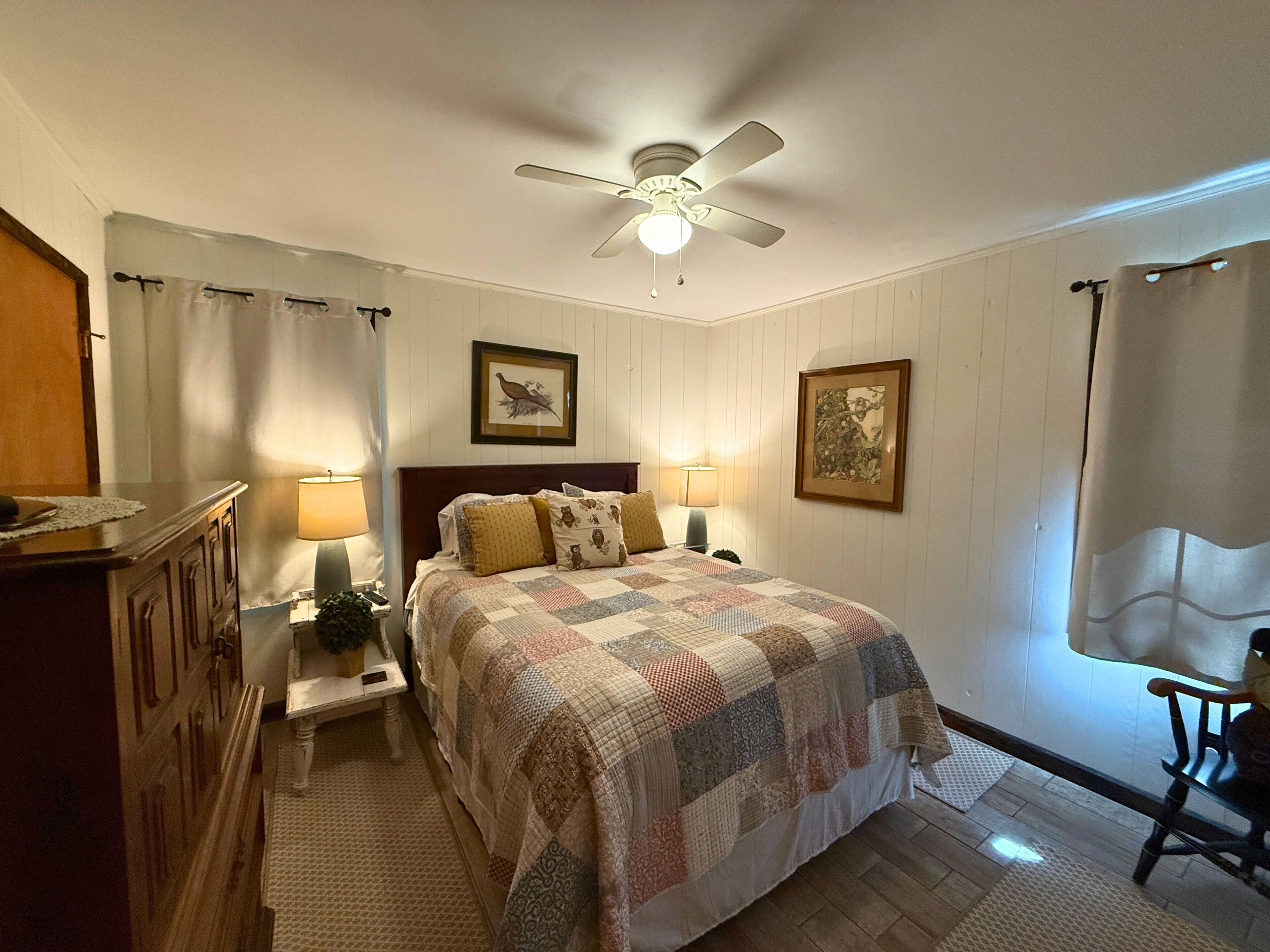 Un dormitorio con cama, dos mesitas de noche con lámparas, un ventilador de techo y dos cuadros en la pared.
