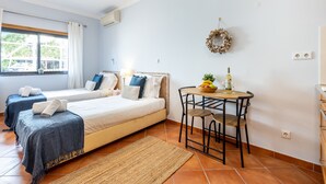 Hotellihuone, jossa on kaksi sänkyä, pieni pöytä, jonka päällä on hedelmäkulho ja pullo, ikkuna, jossa on verhot, ja seinälle ripustettu koriste.