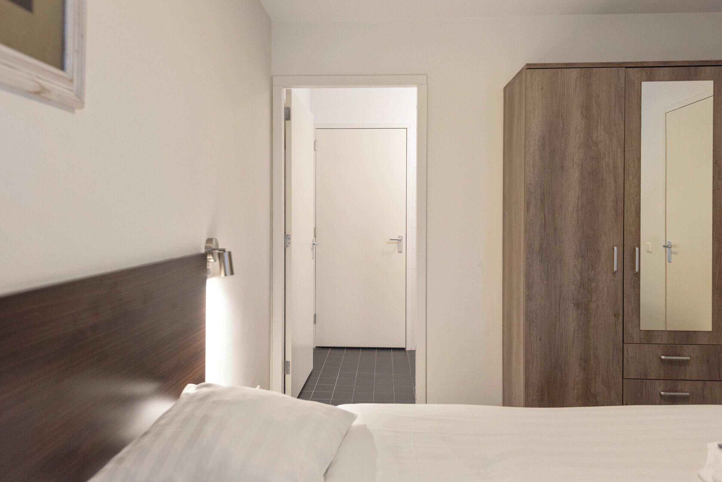 Une chambre d’hôtel avec une tête de lit en bois, un lit avec du linge de maison blanc, une armoire et une porte donnant sur une autre pièce.