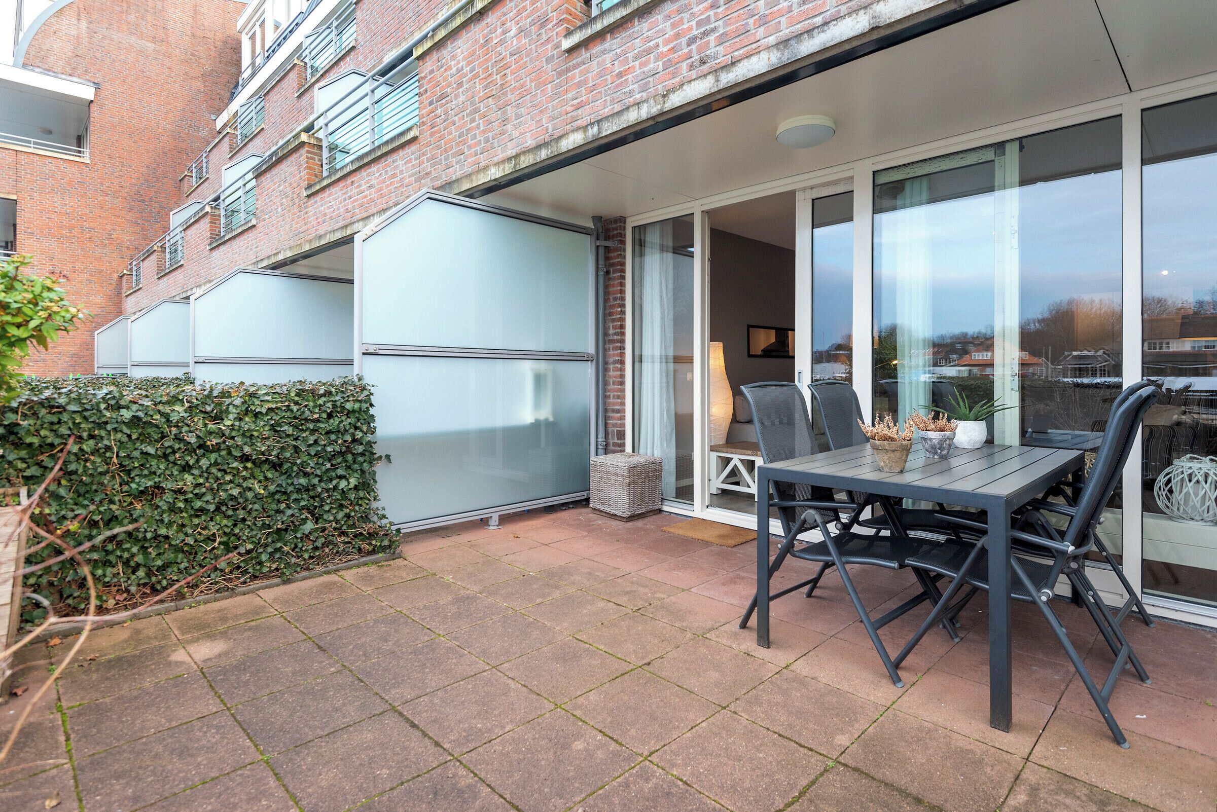 A patio area with a table and chairs, a hedge, and a brick building with a sliding glass door.