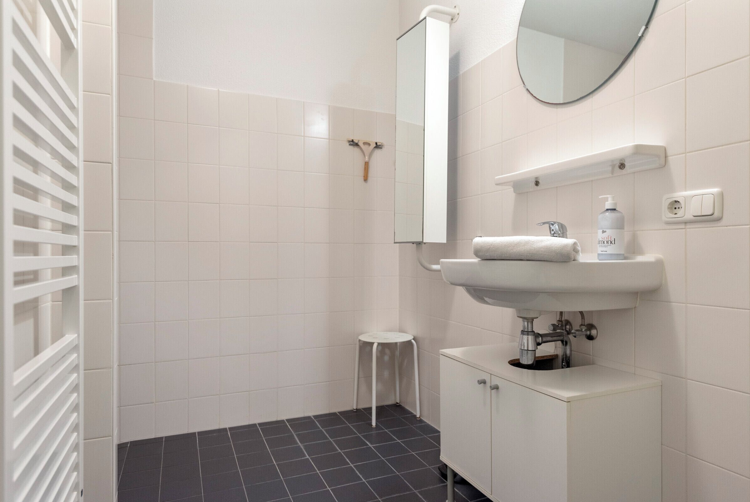 Une salle de bain moderne avec des carreaux blancs, un lavabo blanc, un miroir et un petit tabouret.