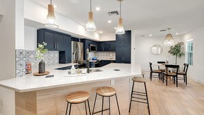 A modern kitchen with dark cabinets, a large island with stools, pendant lighting, and a dining area with chairs. - Palm Beach - Newly Designed come relax, shop, enjoy! (West Palm Beach)