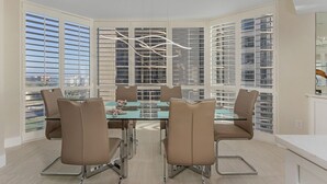 Uma sala de jantar moderna com mesa de vidro e cadeiras marrons, janelas amplas com persianas e vista para edifícios.