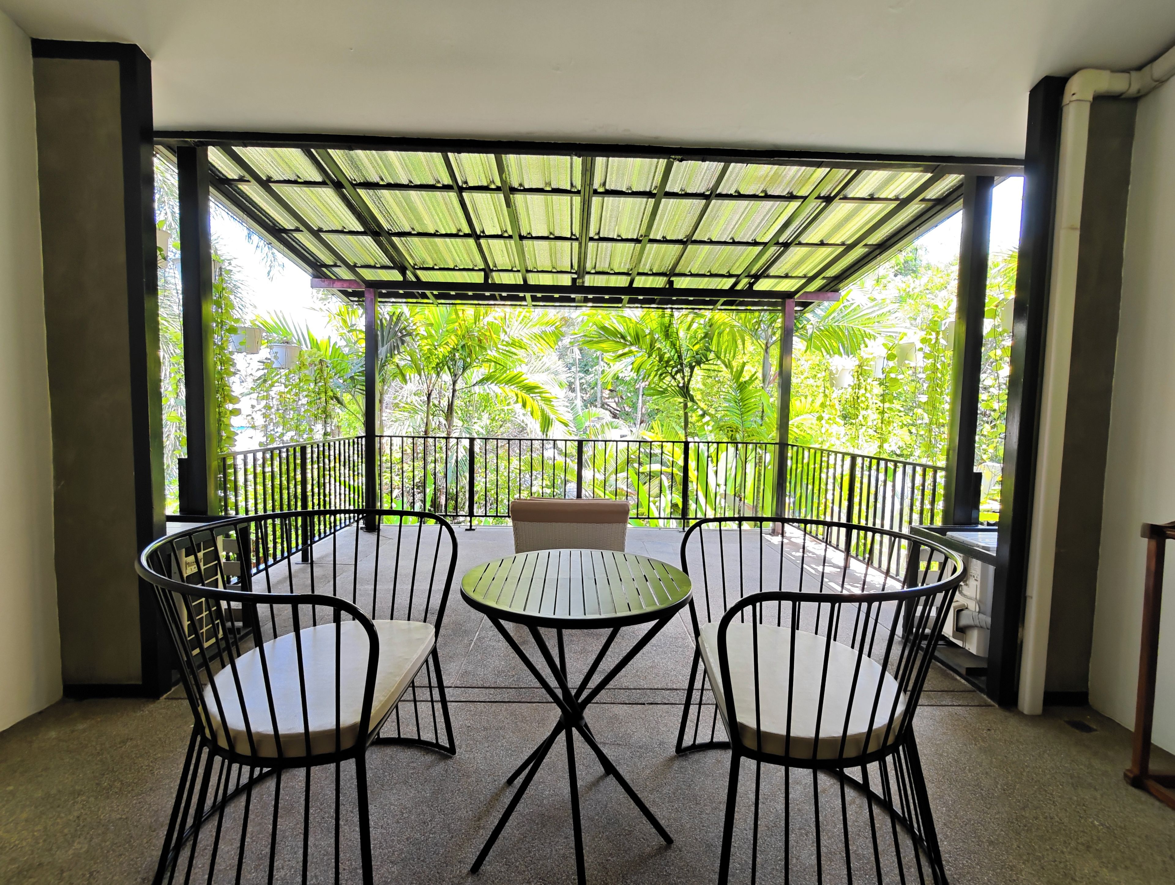 A covered outdoor seating area with a table and chairs, surrounded by a metal railing, overlooking a lush garden.