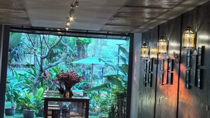 A modern interior with a ceiling fan, a wooden reception desk, and a view of a lush garden through large windows. - Sannot Riverside Boutique Hotel (Chiang Mai)