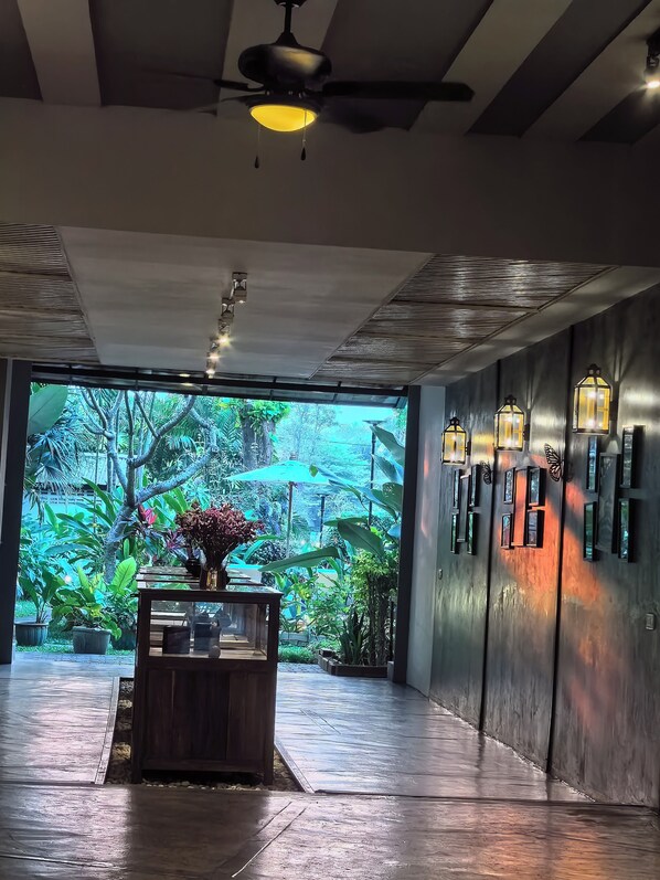 A modern interior with a ceiling fan, a wooden reception desk, and a view of a lush garden through large windows. - Sannot Riverside Boutique Hotel (Chiang Mai)