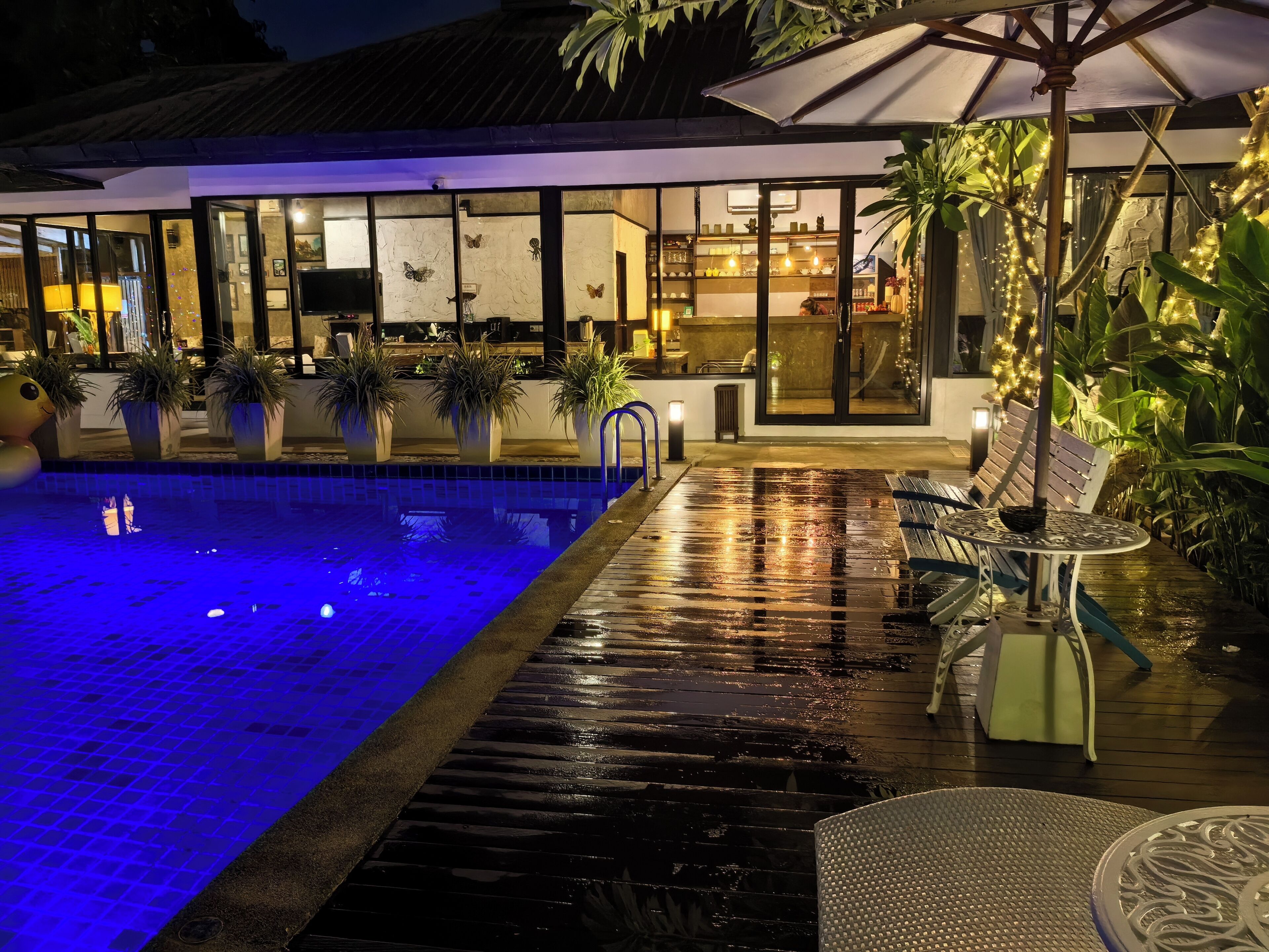 An outdoor pool area with a tiled floor, potted plants, and a seating area with a table and chairs.