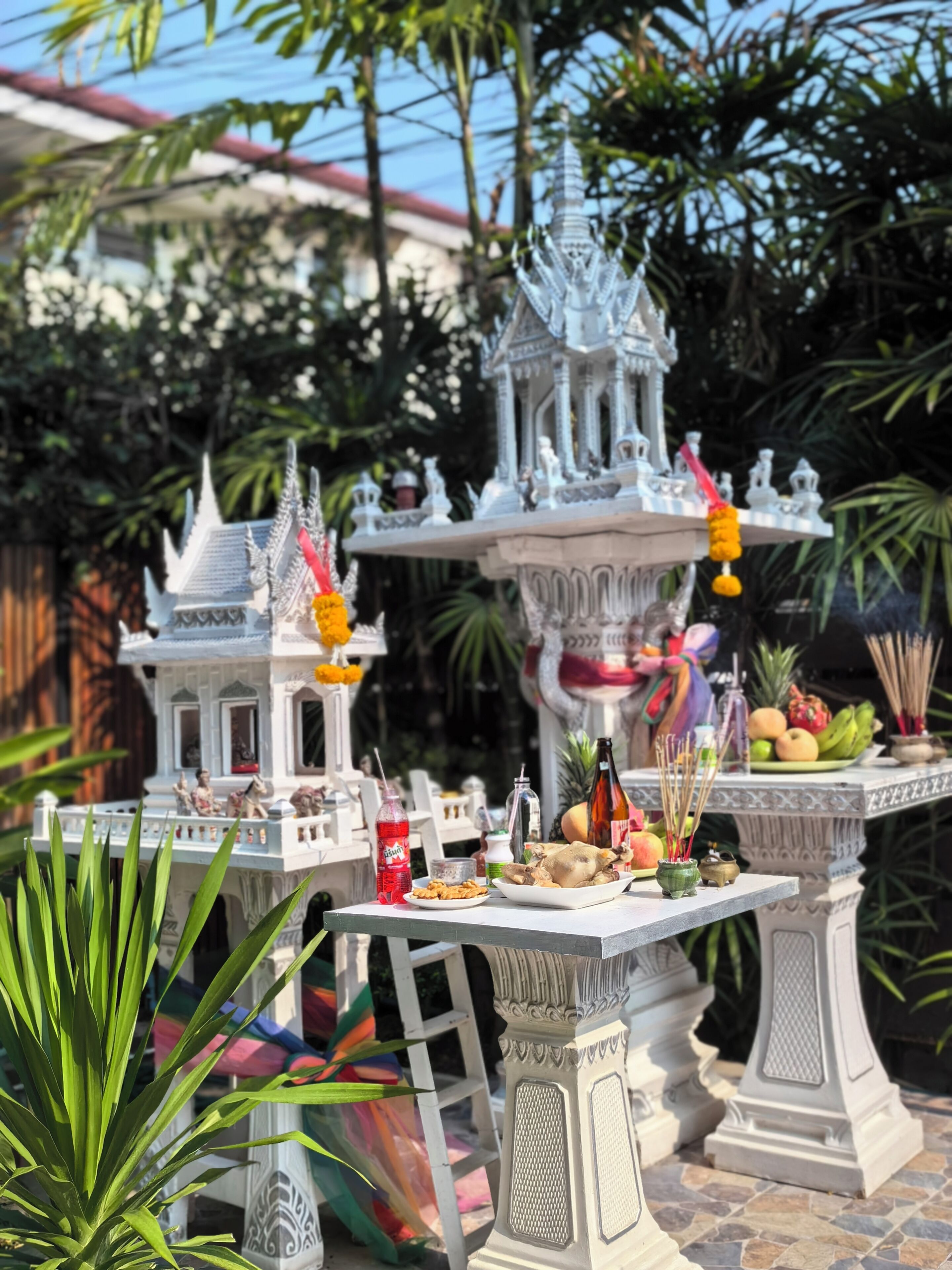 A traditional Thai temple model with intricate architecture, surrounded by lush greenery and a small table with food and incense sticks.