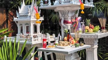 A traditional Thai temple model with intricate architecture, surrounded by lush greenery and a small table with food and incense sticks.