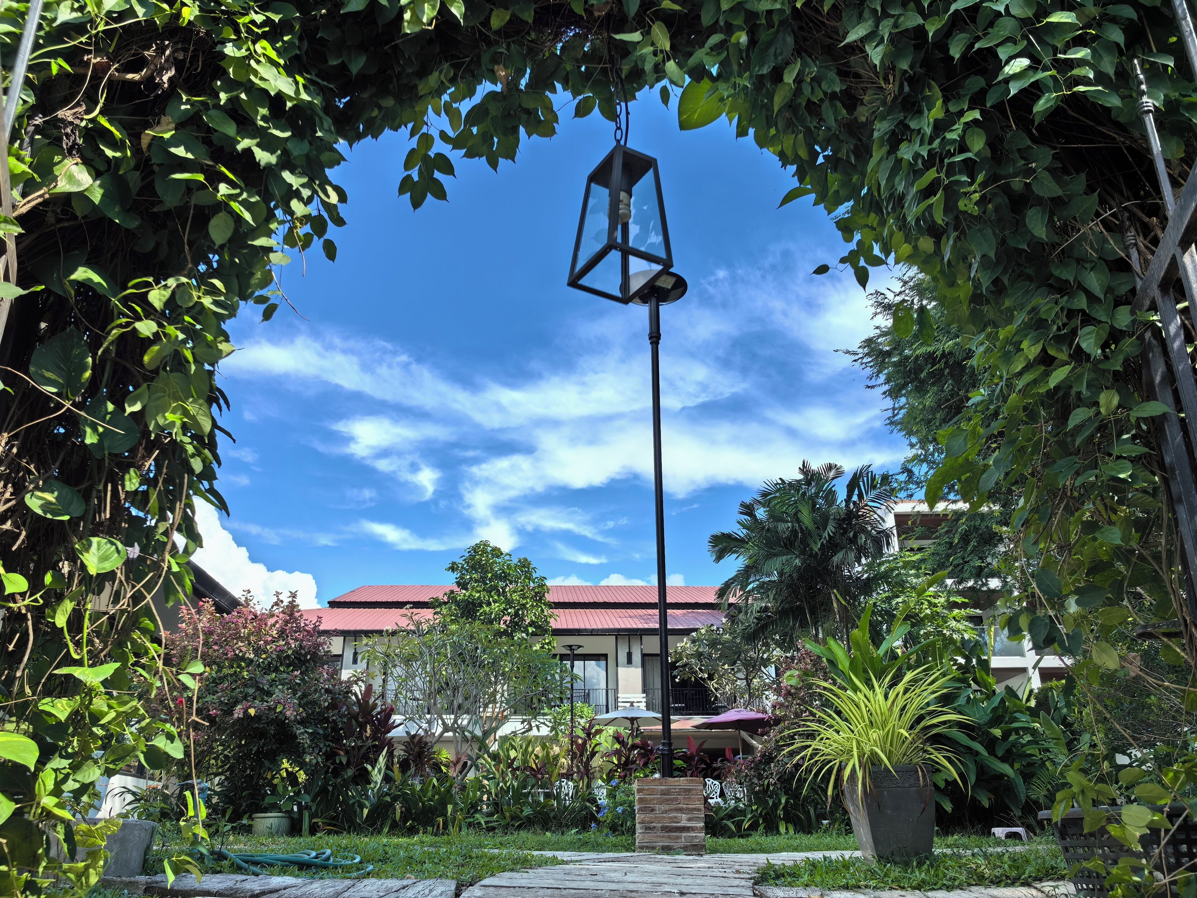 A garden with a pathway, a hanging lantern, and a building with a red roof in the background.