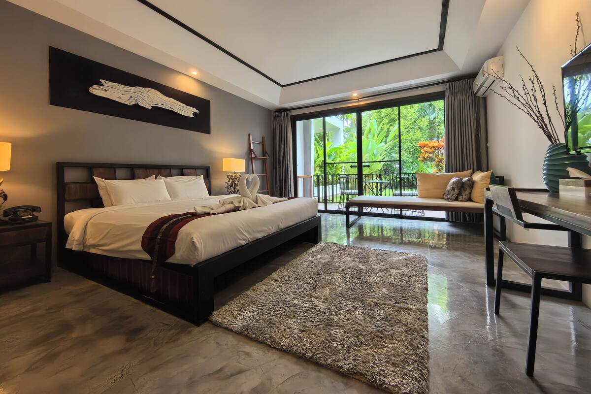 A modern hotel room with a large bed, a desk, and a balcony with a view of greenery.