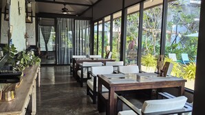 A restaurant with a long counter, multiple tables, and chairs. There are ceiling fans, large windows with curtains, and a view of greenery outside. - Sannot Riverside Boutique Hotel (Chiang Mai)