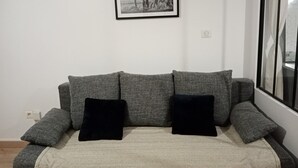 En grå soffa med två svarta kuddar, ett inramat svartvitt fotografi på väggen och ett fönster med gardiner.