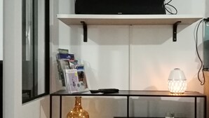 Ett vardagsrum med en svart tv på en vit vägg, en svart möbel för underhållningsutrustning med en vas och tidskrifter, och en tänd dekorativ lampa.
