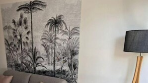 A modern living room with a grey sofa, a coffee table with a fruit bowl, and a wall mural of tropical trees. - Ô Lagon Accès Direct Plage (LA SALINE LES BAINS)