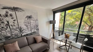 A modern living room with a grey sofa, a dining table with chairs, a large window with a view of greenery, and a wall with a black and white mural. - Ô Lagon Accès Direct Plage (LA SALINE LES BAINS)