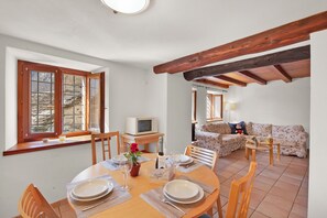 A dining area with a wooden table set for four, a sofa, and a television in the background. - Rustico Esterina (Sonogno)