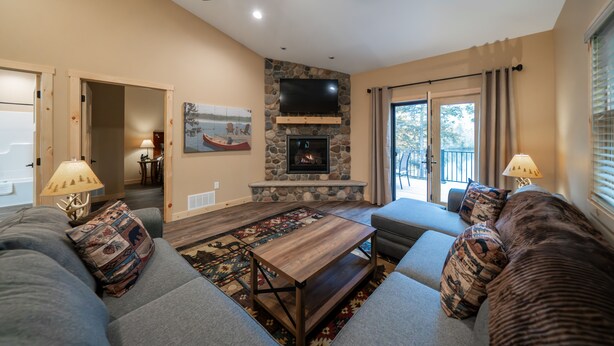 A living room with a stone fireplace, a ceiling fan, and a large window with curtains. - New! 4BR Lake Namakagon Cabin - Sleeps 15 - Lakefront w/ boat access (Cable)