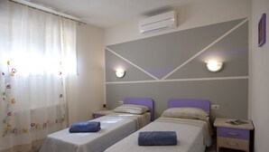 A hotel room with two beds, each with a blue towel, a nightstand, a wall-mounted air conditioner, and a decorative curtain with floral patterns. - Borgo dell'Uccellaia - Smeraldo (Marciana Marina)