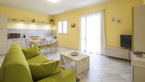 A bright living room with a green sofa, a dining area with chairs, a kitchen with a microwave, and a TV. - Borgo dell'Uccellaia - Smeraldo (Marciana Marina)