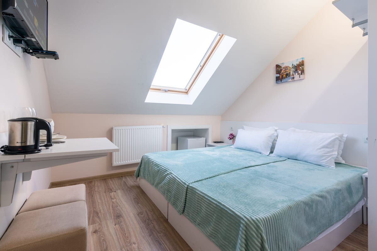 Un dormitorio con tragaluz, cama con colcha verde, mesita de noche con lámpara y radiador.