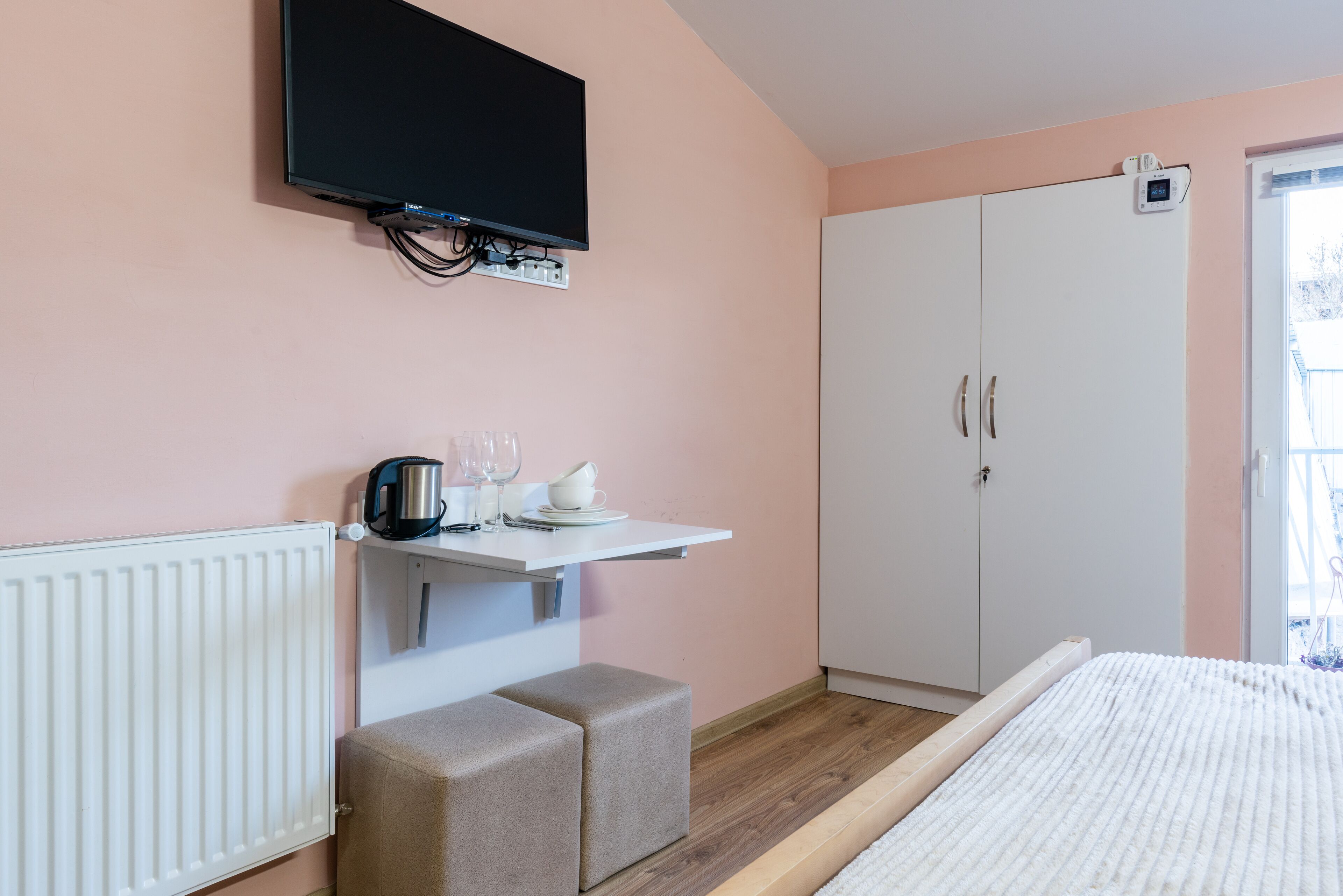 A bedroom with a bed, a white wardrobe, a television mounted on the wall, a small table with a kettle, cups, and a plate, and a window with a view of the outside.