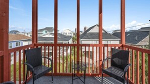 A covered balcony with two wicker chairs and a small table, overlooking a residential area with houses and a clear blue sky.