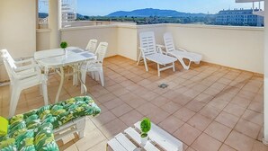 Un balcón con una mesa, sillas, una planta pequeña y vistas a montañas distantes.
