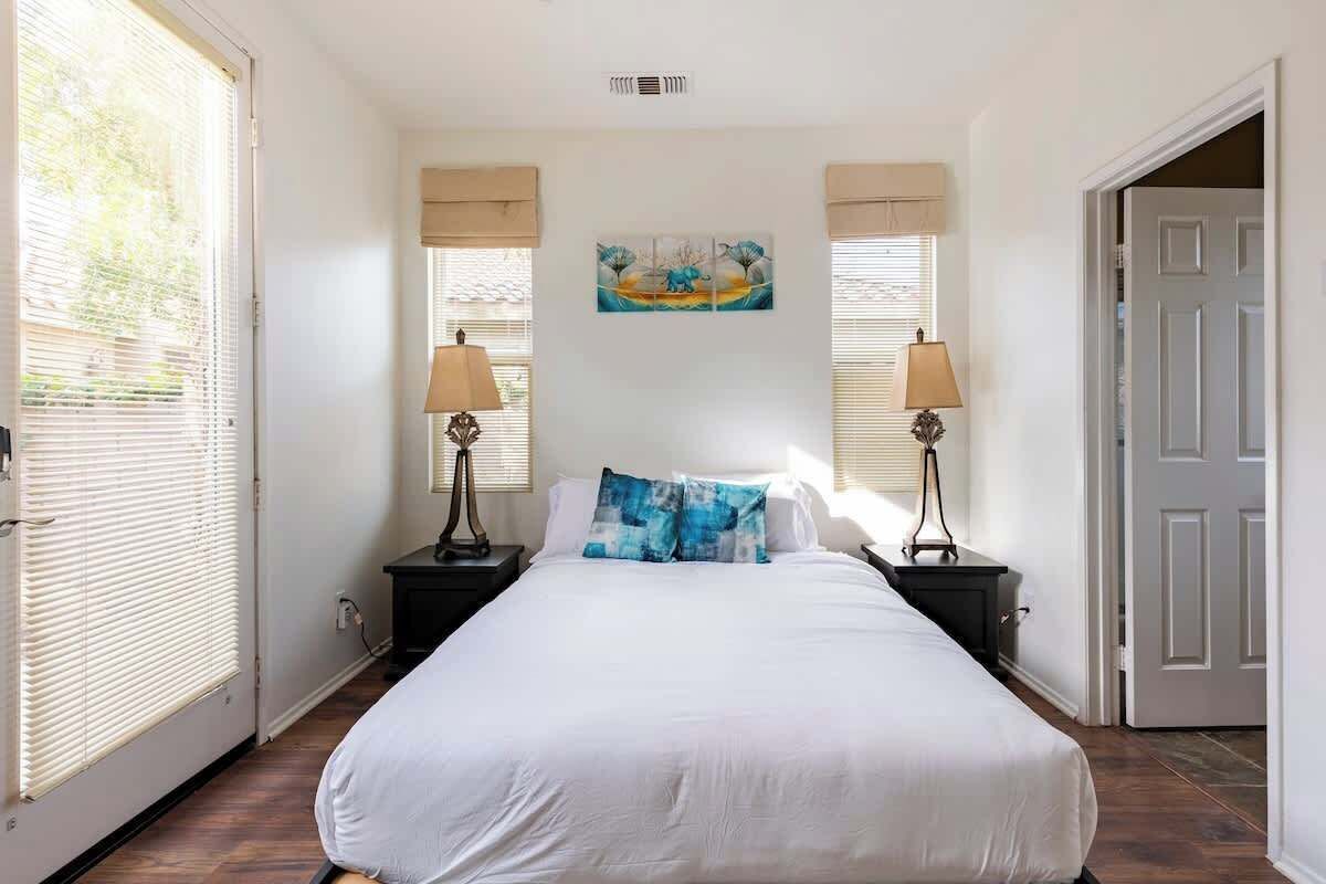 A bedroom with a large bed, two bedside tables with lamps, a painting on the wall, and a door to the right.
