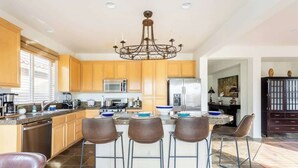 A modern kitchen with wooden cabinets, a central island with bar stools, stainless steel appliances, and a chandelier. - Cozy Home in Gated Country Club (Indio)