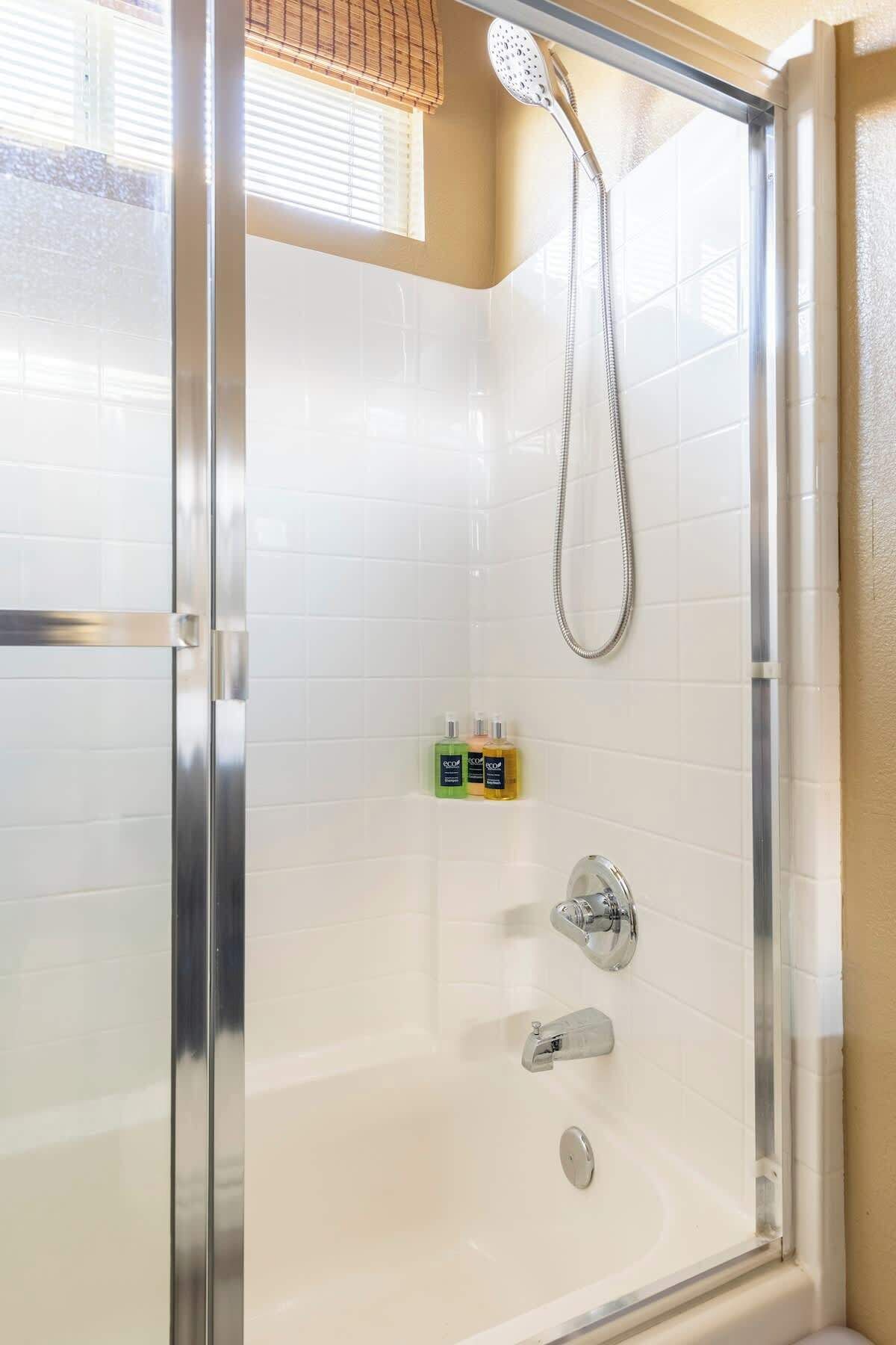 Una cabina doccia in vetro con soffione a mano, una vasca da bagno e due flaconi di shampoo appoggiati al muro.