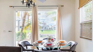 A dining area with a glass table, black leather chairs, and a view of a house and palm trees through the window. - Cozy Home in Gated Country Club (Indio)