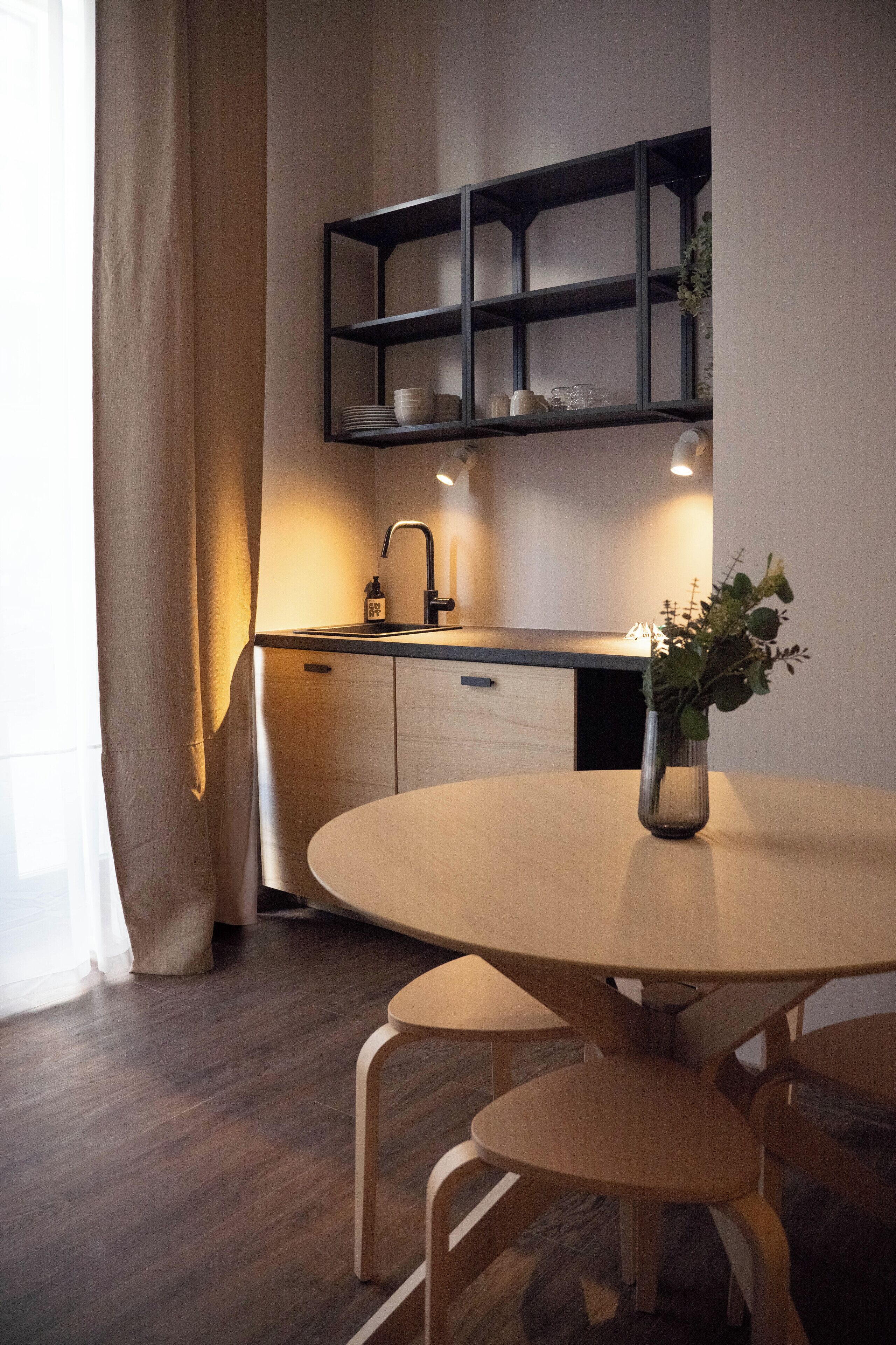 A modern kitchen with a round wooden table, a black shelving unit above the counter, and a vase with greenery on the table.