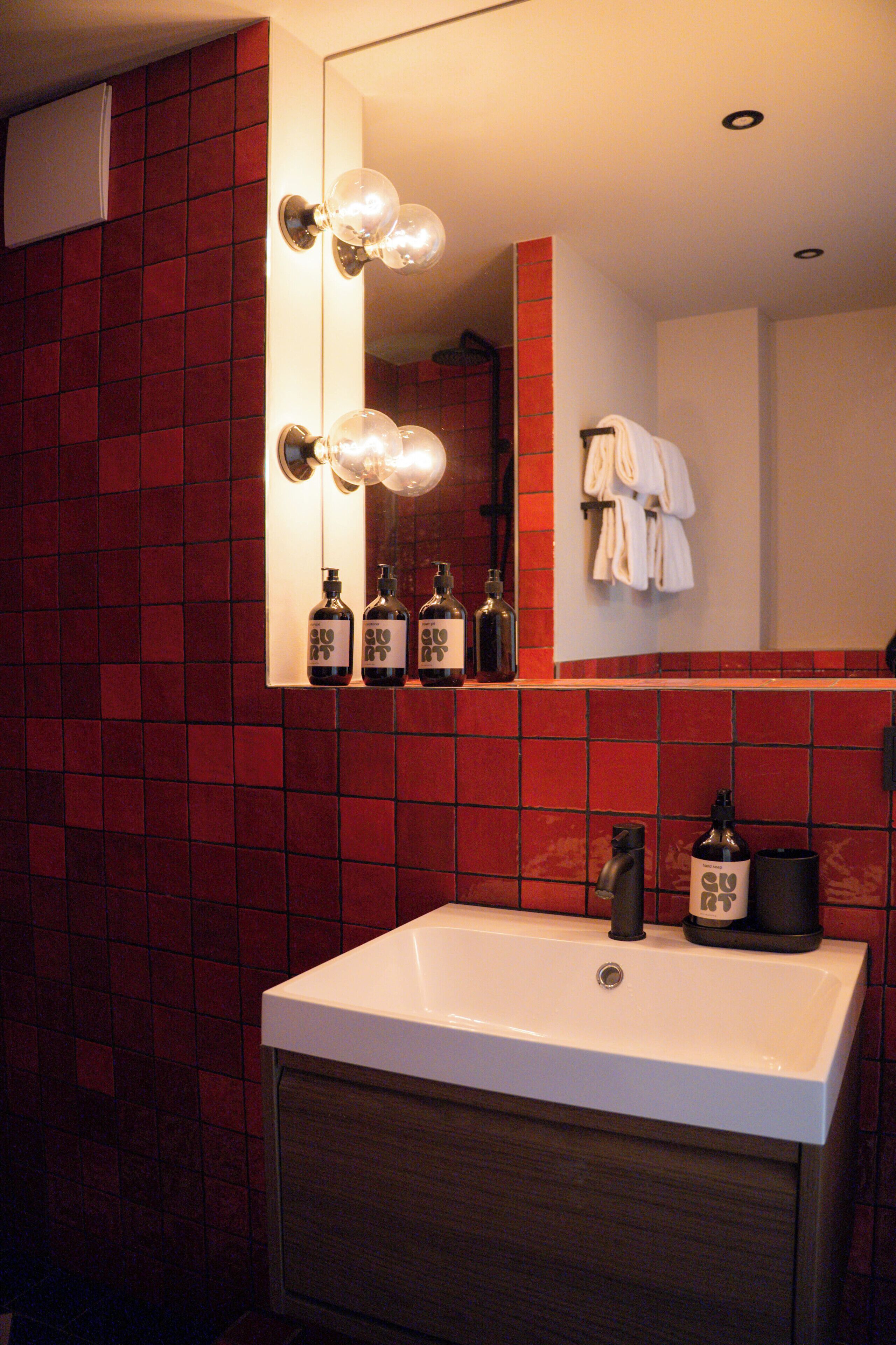 Un baño con lavamanos blanco, paredes de baldosas rojas, un espejo con luces y un perchero para toallas.