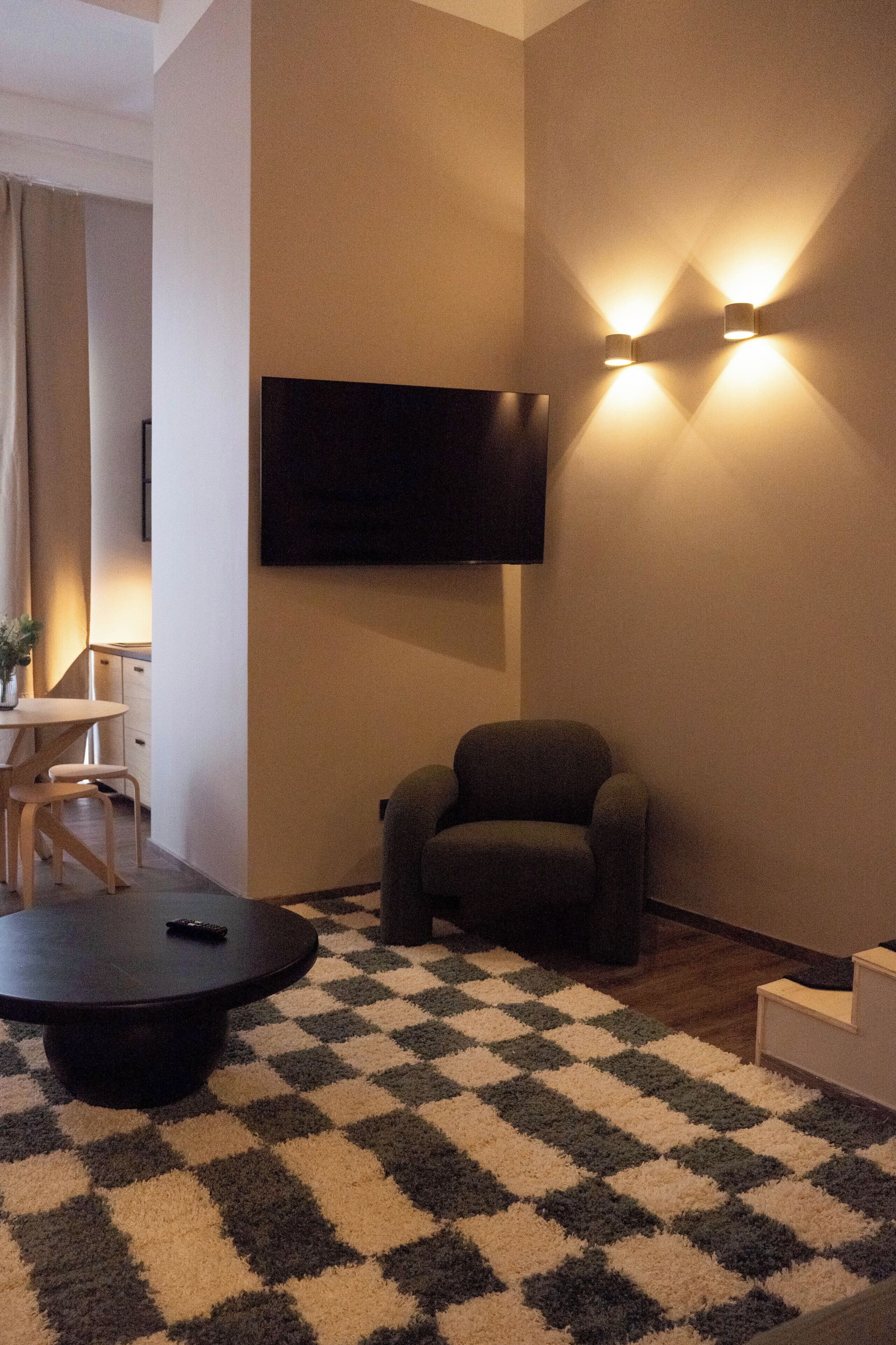 Un salon avec un téléviseur à écran plat fixé au mur, un tapis à carreaux, une table basse ronde et un fauteuil.