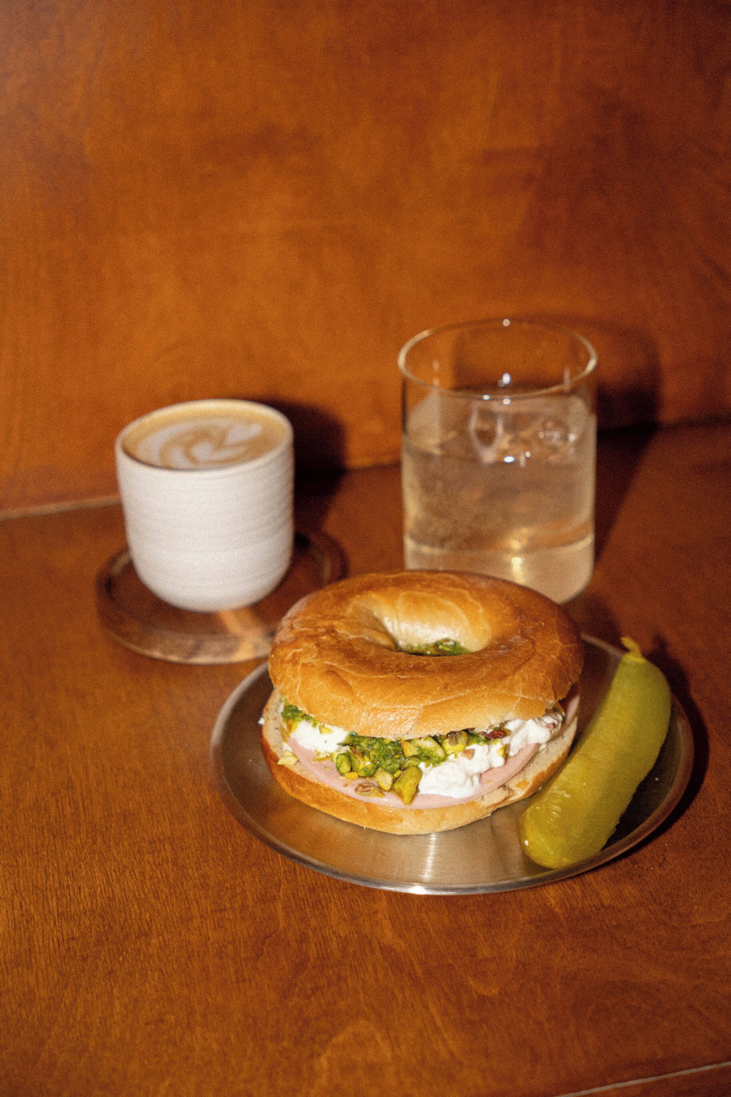 Un sándwich en un plato con un pepinillo, una taza de café y un vaso de agua sobre una mesa de madera.