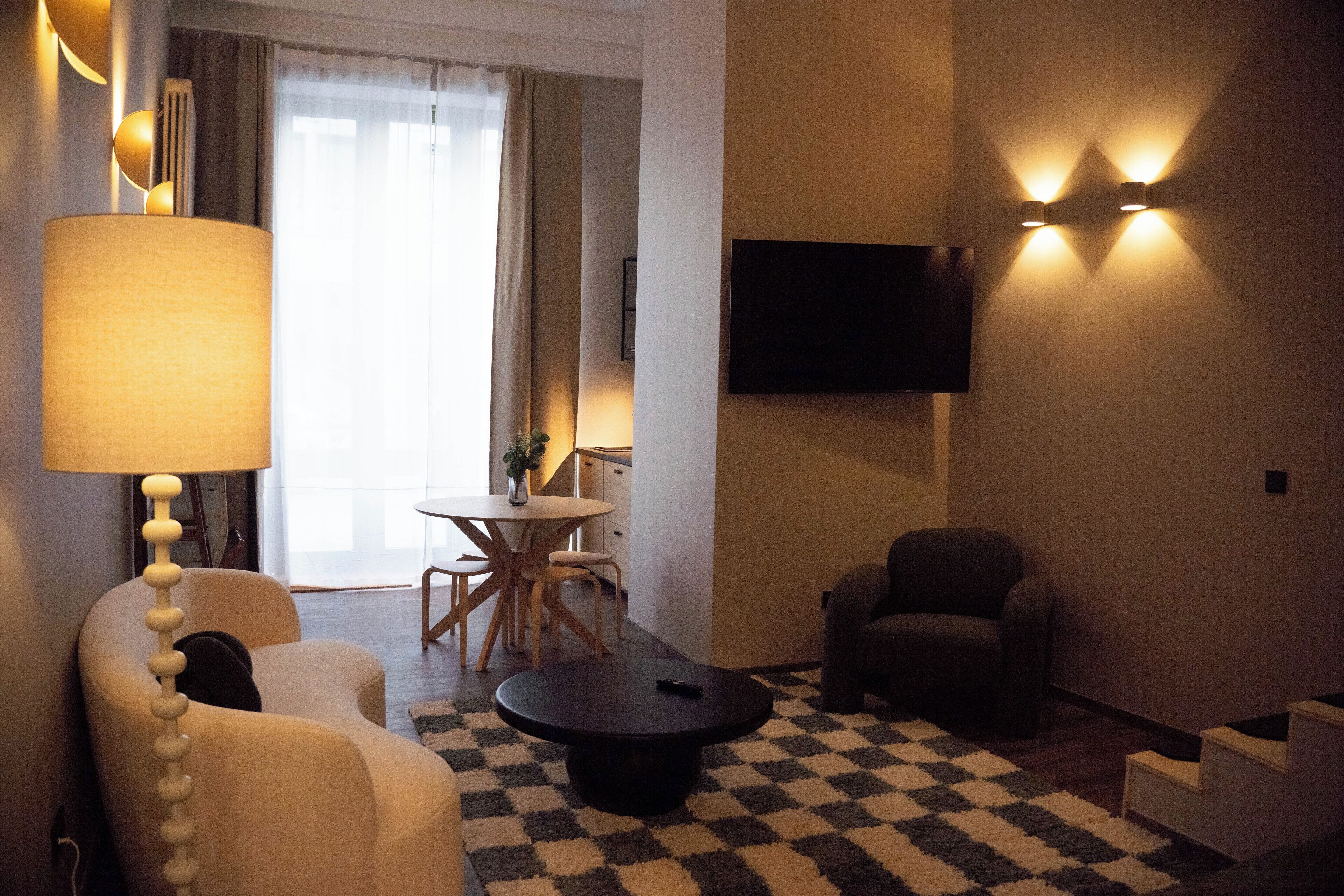 Ein Wohnzimmer mit Sofa, Sessel und Couchtisch. Ein Fernseher ist an der Wand montiert und links steht eine Lampe.
