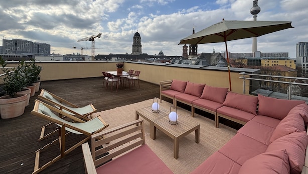 A rooftop terrace with a wooden deck, red seating, and a table with a lamp. A large umbrella provides shade. - The Charming by Curt Suites (Berlin)