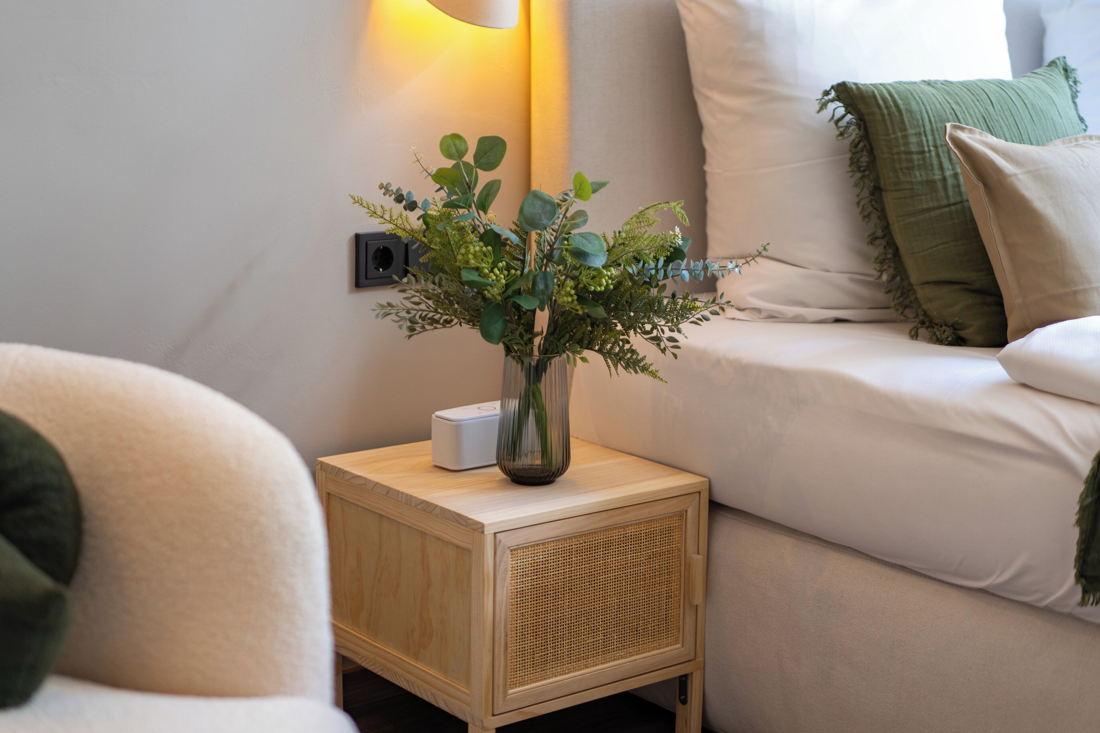Une petite table de chevet en bois, sur laquelle se trouve un vase rempli de verdure et un objet blanc, placée à côté d’un canapé beige orné d’un coussin vert.
