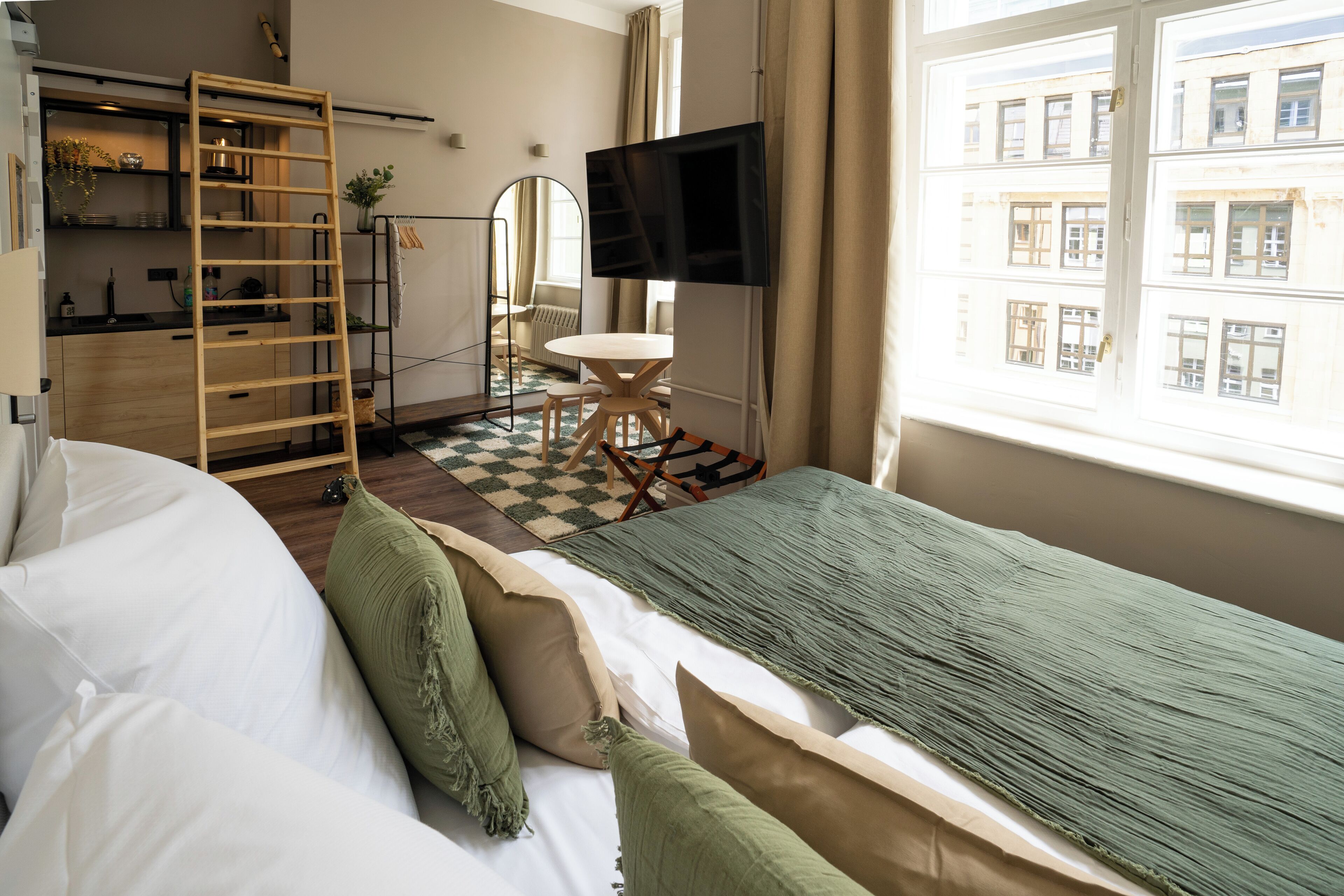 Een moderne hotelkamer met een bed, een televisie, een bureau met stoel, een ladder en een raam met uitzicht op gebouwen.