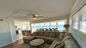 A spacious living room with a large sectional sofa, a small round table, and a view of the ocean through large windows. - Beachfront in Surfside Shores (Gulf Shores)