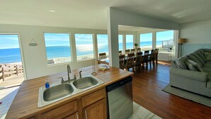 A modern kitchen with a dishwasher, double sink, and wooden cabinets. There is a dining area with a table and chairs, and large windows offering a view of the beach. - Beachfront in Surfside Shores (Gulf Shores)