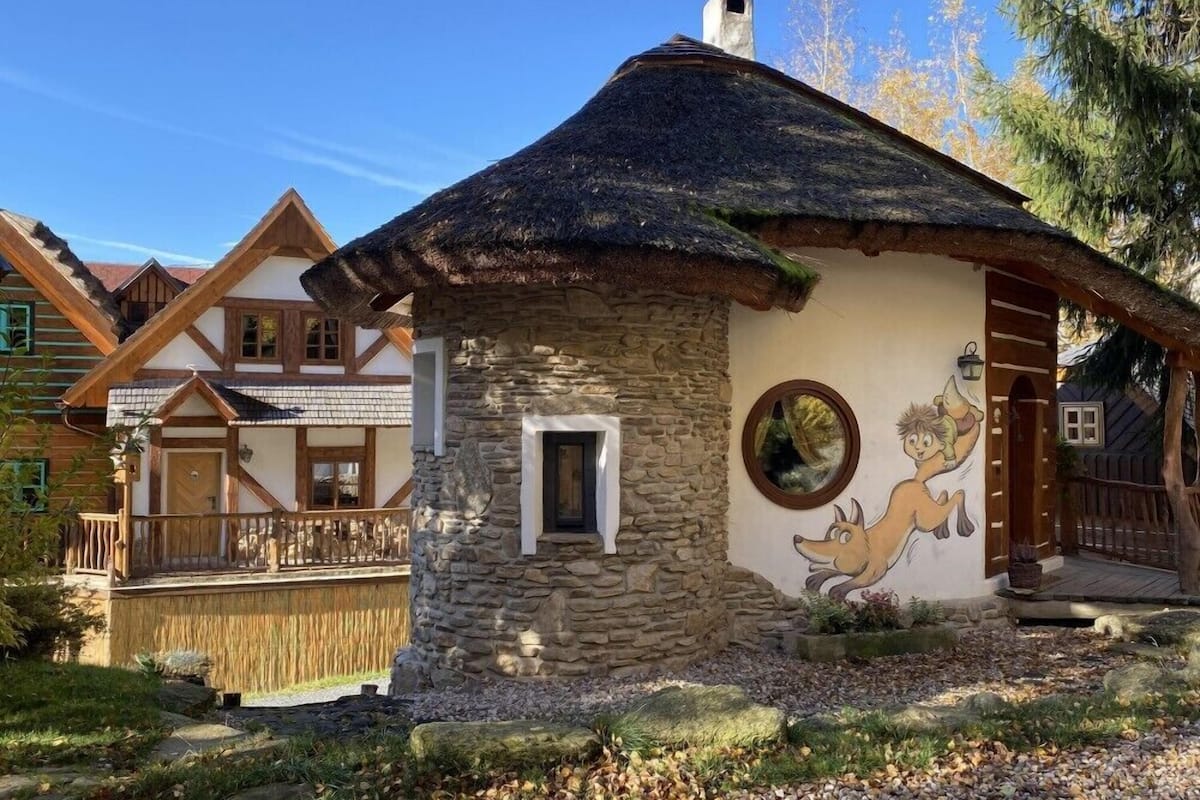 Una casa rústica con pared de piedra y techo de paja, rodeada por una valla de madera y un jardín.