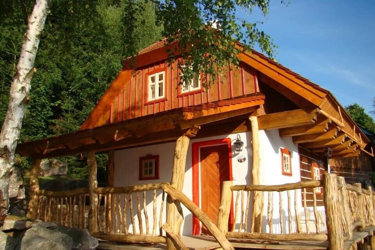Una cabaña rústica con porche de madera y puerta roja, rodeada de árboles y rocas.