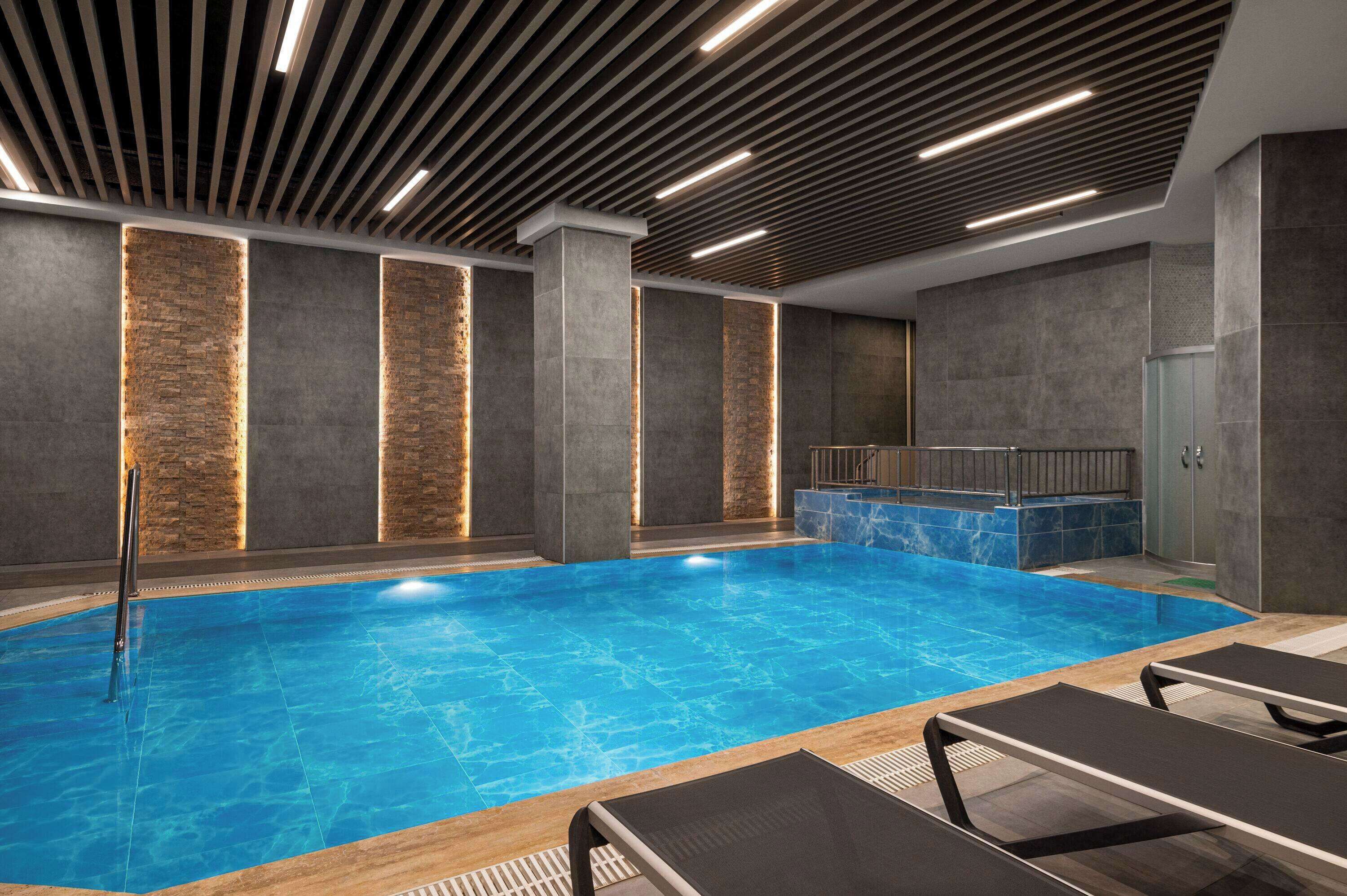 Una piscina interior moderna con superficie de agua azul cristalina, rodeada de sillones de descanso y una pared de piedra con iluminación empotrada.