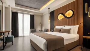 Habitación de hotel moderna con una cama grande, un escritorio y una silla. Cuenta con una ventana con cortinas, una lámpara fijada a la pared y un diseño de paneles de madera en la pared.