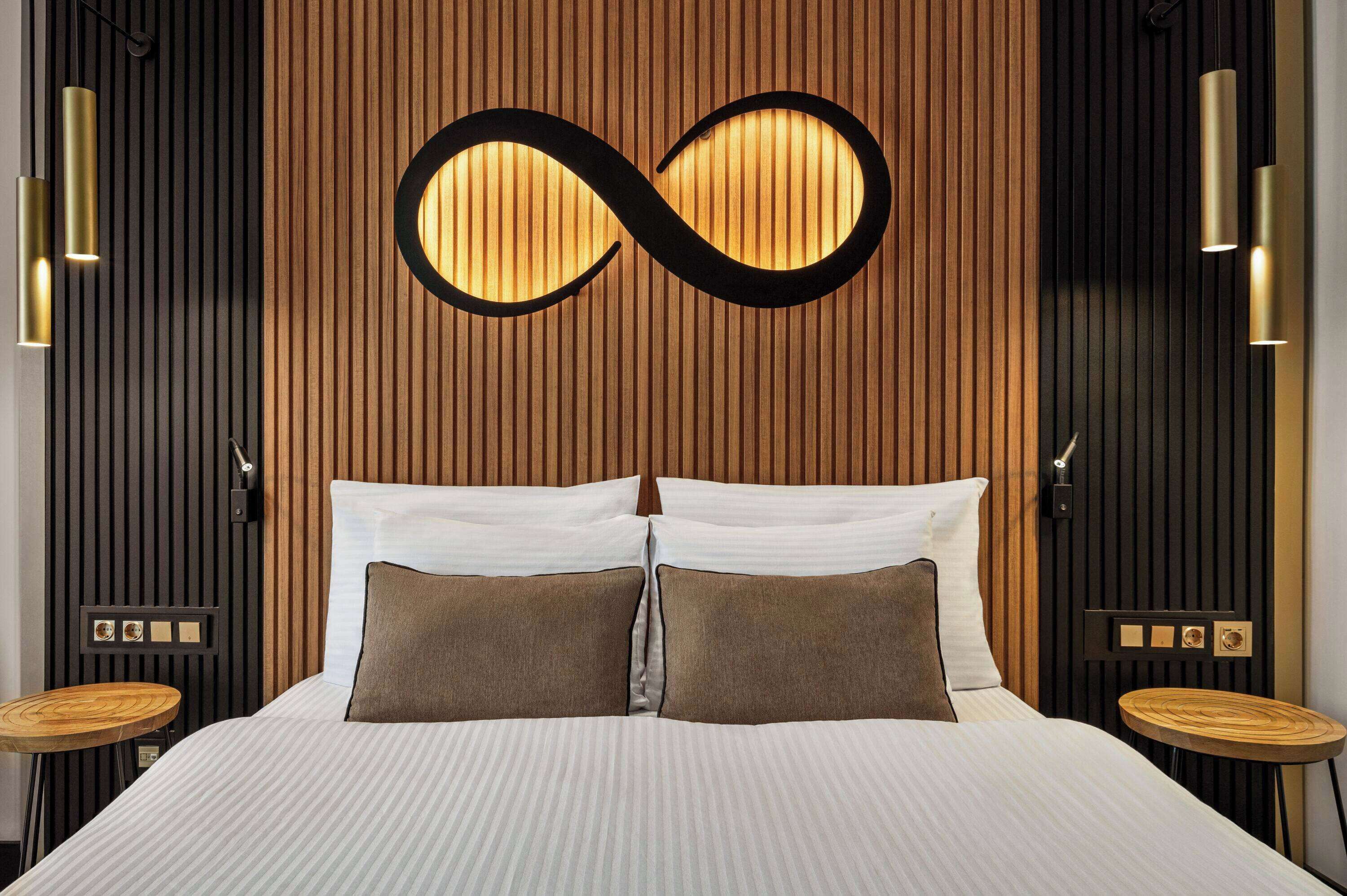 A modern hotel room with a bed, two bedside tables, a wall-mounted infinity mirror, and vertical wooden paneling.